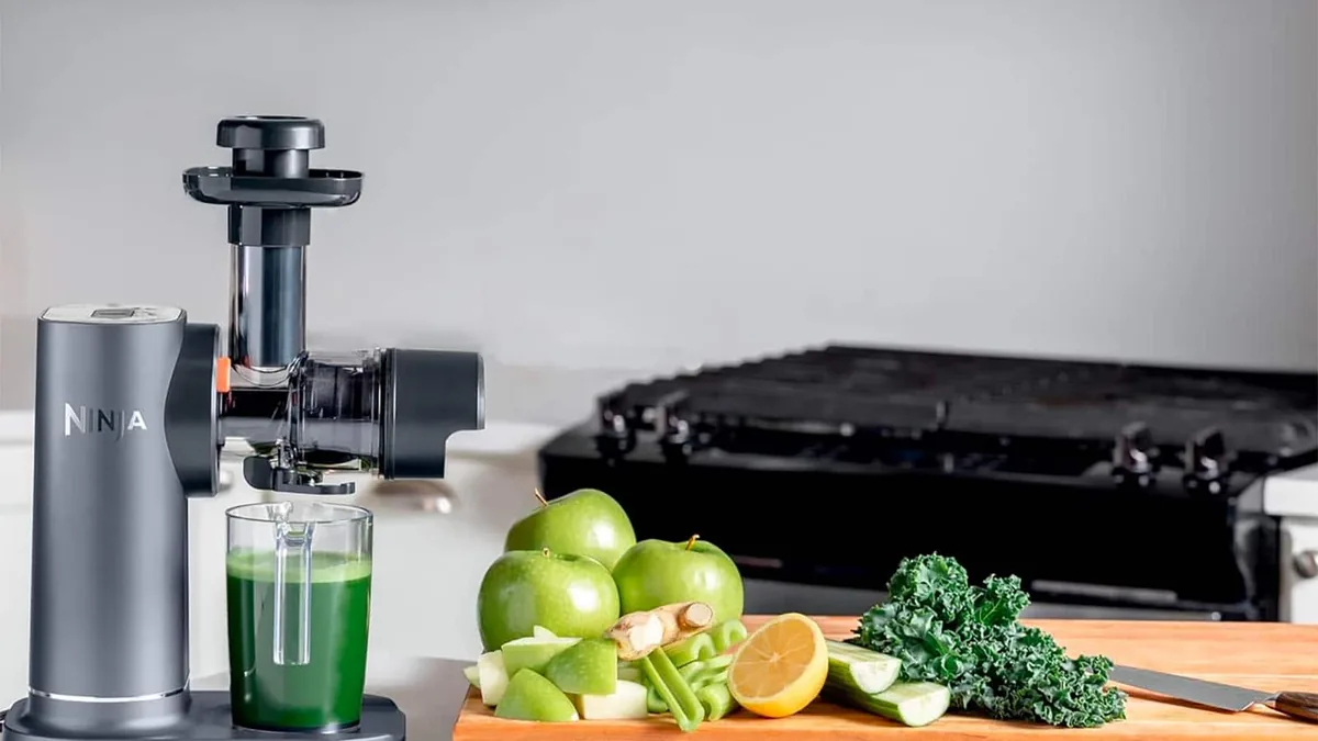 Extracteur de jus gris avec jus vert et planche de decoupe garnie de pommes vertes, kale et gingembre