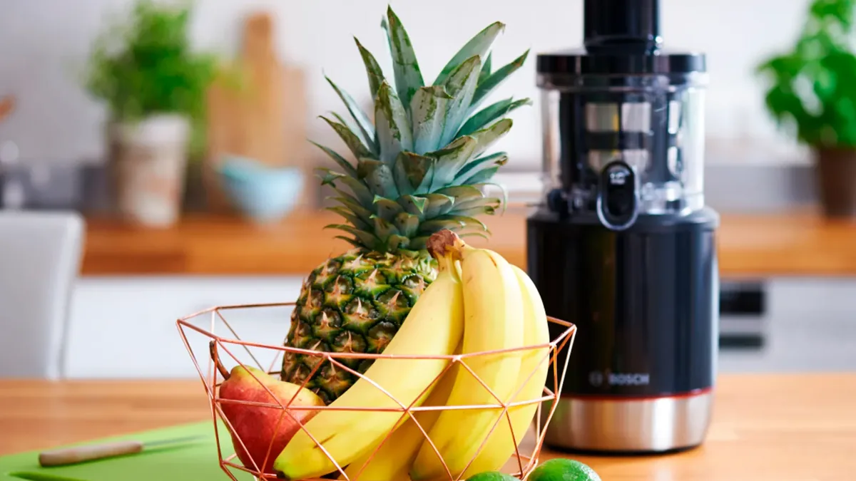 Extracteur de jus noir pose pres d'un panier d'ananas, bananes et citrons verts sur plan de travail bois